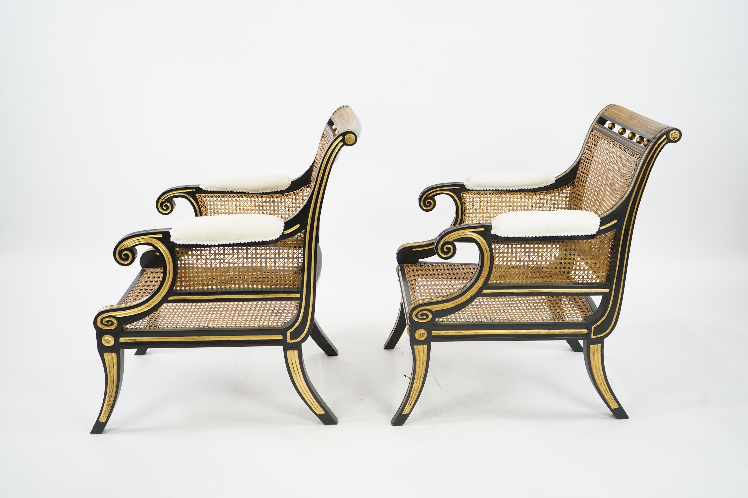 A pair of Regency style ebonised, parcel gilt caned bergere elbow chairs